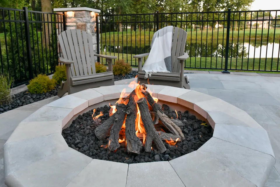 Close view of round firepit, poolside chairs, paver deck, fence, and planting